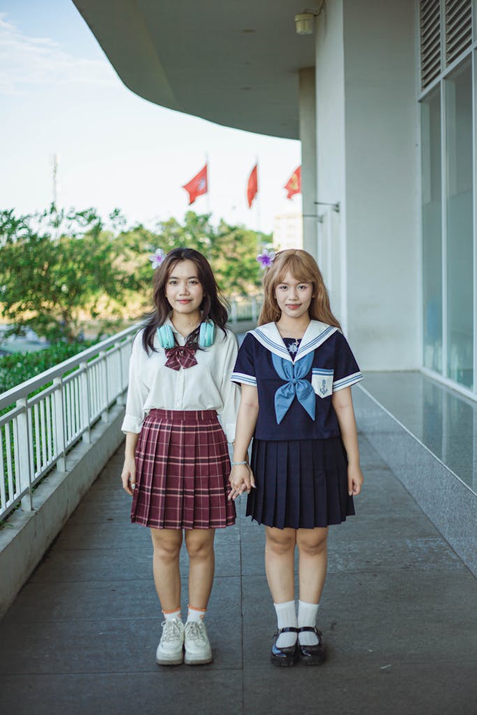 Two young girls holding hands in school uniforms, standing outdoors by a modern building.